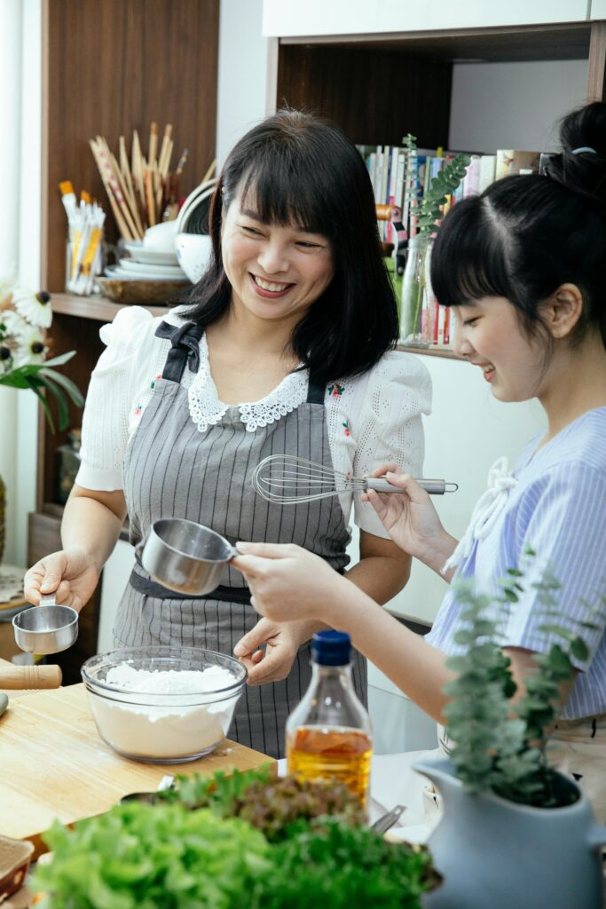 Adult happy Asian lady wearing apron standing near wooden table and discussing with teenage daughter details of recipe while cooking together in kitchen
